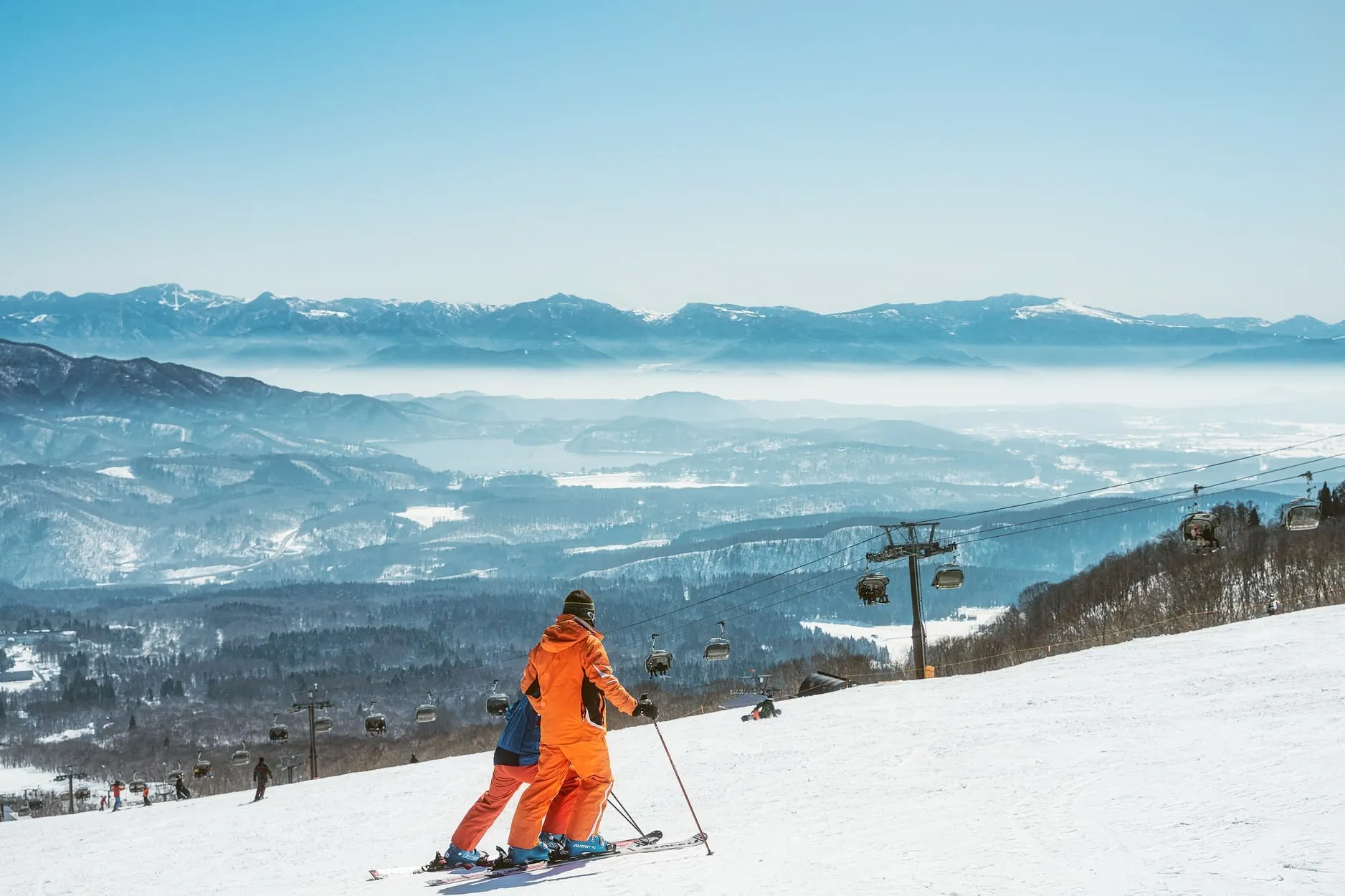 Skiers descending a wide groomed slope with chairlifts and snowy mountain ranges in the distance